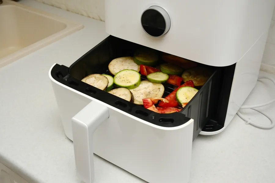 Airfryer täynnä viipaloituja munakoisoja ja kesäkurpitsaa, valmisteilla maukas kasvisateria.