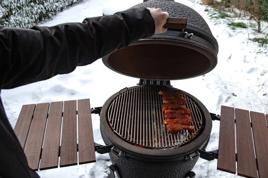 Henkilö avaa grilliä, jossa on ribsejä, luminen maisema taustalla. Grilli on pyöreä ja hieman savinen, ja lähistöllä on puinen työtaso.