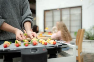 Mies grillaa kasvisvartaita sähkögrillillä ulkona, taustalla ystäviä nauttimassa kesästä.